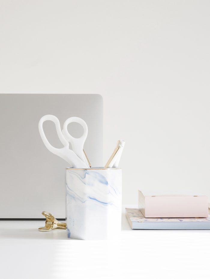 Desktop with silver laptop, white and blue cup holding white scissors and pens, and stacked notebooks