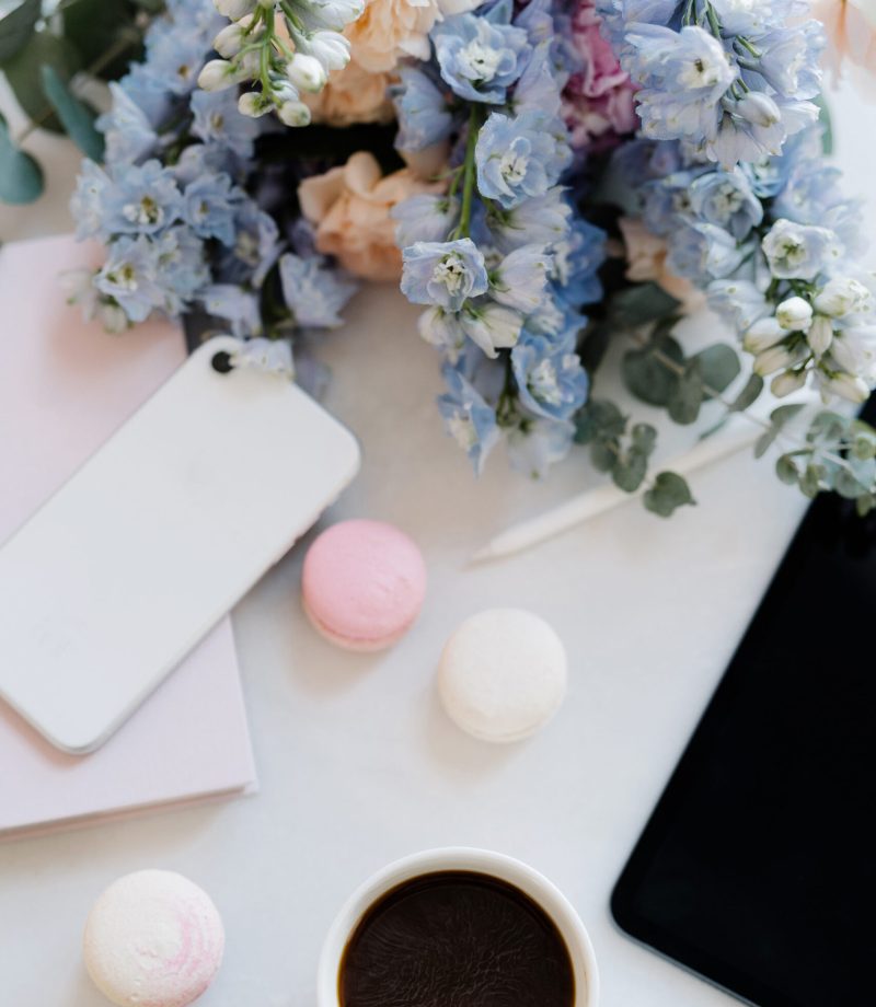 Pastel colored flowers on white desktop. Blank Ipad, cup of coffee, macarons, white Iphone and pink book